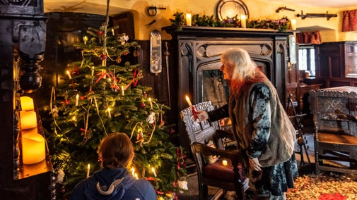 Warmly lit room with a Christmas tree; one person holds a candle near another seated by the tree.